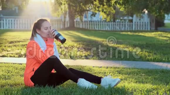 年轻漂亮的女人穿着红色运动衫在晨跑后坐在草地上视频的预览图