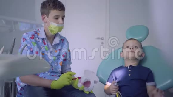 十几岁的男孩戴着医用口罩和手套小孩子在牙医办公室玩耍年轻人教孩子刷牙视频的预览图
