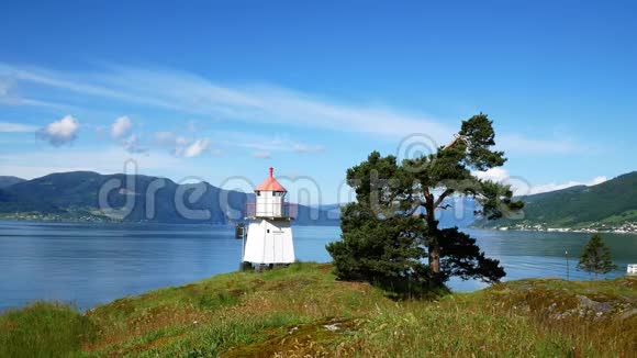 在一个阳光明媚的日子里白色灯塔塔上有红色的顶部和附近的常青树沿着挪威峡湾两岸的山视频的预览图