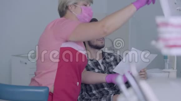 戴着粉红色面具和手套的女牙医在屏幕上向男性病人展示他牙齿的照片年轻人视频的预览图