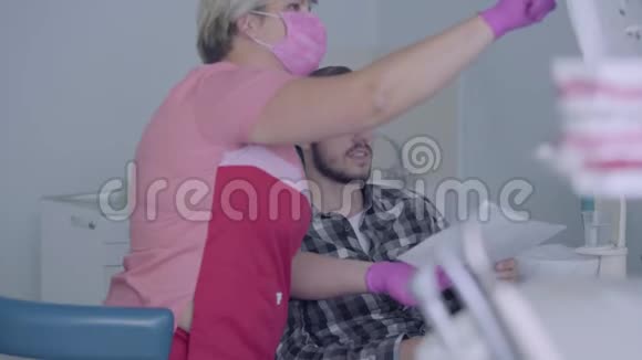 戴着粉红色面具和手套的女牙医在屏幕上向男性病人展示他牙齿的照片年轻人视频的预览图