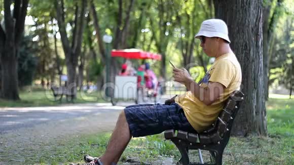 戴帽子戴太阳镜的人正在长凳上的公园里使用智能手机男子使用智能手机在树下的公园放松视频的预览图