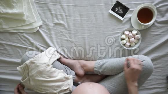 孕妇在床上准备婴儿衣服怀孕和分娩概念的俯视图视频的预览图