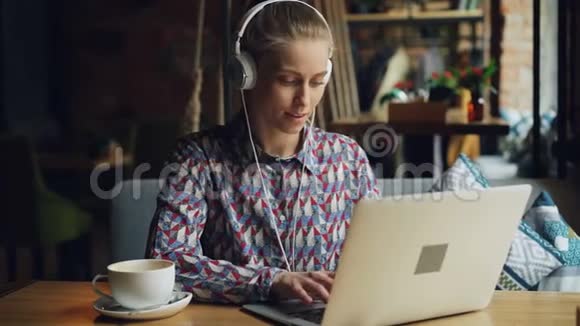 带着耳机听音乐的年轻女性在咖啡馆里用笔记本电脑听音乐视频的预览图