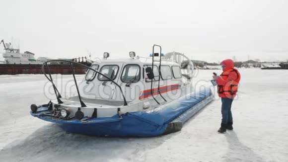 穿橙色夹克的女人在气垫船背景上使用手机冰冻水冬湖上的浮船冬季船只视频的预览图