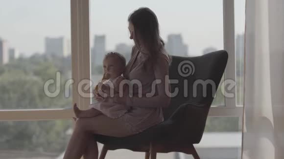年轻漂亮的女人坐在窗前舒适的椅子上怀里抱着女婴一种概念视频的预览图