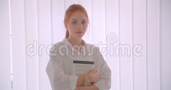 年轻漂亮红头发的女学生举着一本书看着站在白色房间里的照相机的特写照片视频的预览图