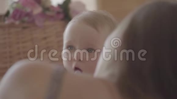 漂亮的女婴喝着瓶子里的水坐在妈妈家附近的床上可爱的白种人在室内视频的预览图