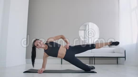 公寓里的黑发女人做一个运动外侧木板站在她的膝盖上抬起她的腿健身视频的预览图