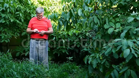 穿休闲服装的老人也许是一个养老金领取者在后院或花园里使用他的智能手机视频的预览图