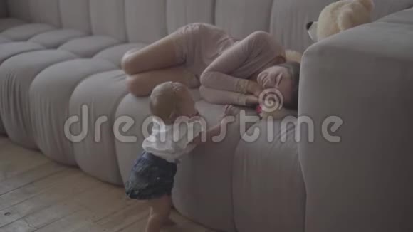 漂亮苗条的年轻女子躺在沙发上休息而她的小宝贝女孩站在附近妈妈在玩视频的预览图