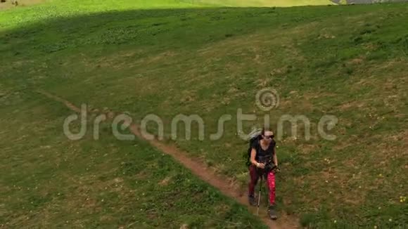 穿着紧身裤的女孩背着背包和徒步旅行杆在绿色的山坡上散步视频的预览图