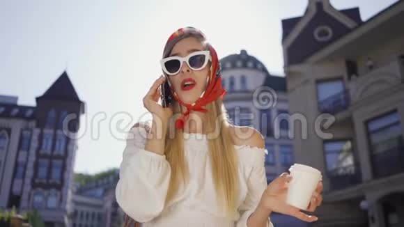 肖像时尚漂亮的年轻女人带着一杯咖啡站在街上晒太阳用手机说话视频的预览图