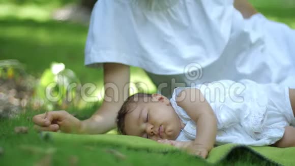 妈妈吻着睡在户外绿草地上的宝宝绿色夏日公园里快乐的年轻母子美丽的视频的预览图