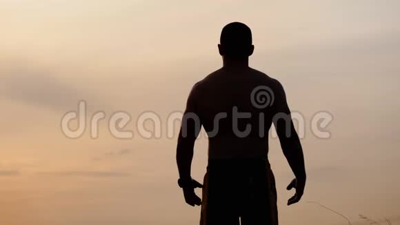 从背部看一个赤裸躯干的运动男子的轮廓显示他的肌肉和二头肌靠在天空视频的预览图