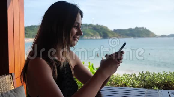 一个年轻的女孩坐在露台上点击电话她甜甜地笑着正在和海滩上的人聊天视频的预览图