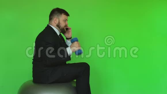 商人手里拿着哑铃坐在健身球上打电话有趣的情况铬绿色有胡子的视频的预览图