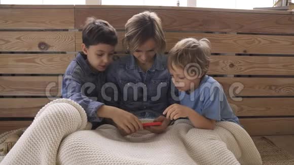 年轻的高加索女人有两个孩子玩智能手机在沙发上放松快乐的男孩喜欢在家里度过时光视频的预览图