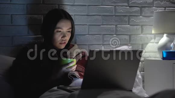 女孩感到恶心躺在床上喝茶在笔记本电脑上看电影流感季节视频的预览图