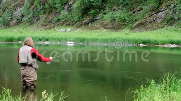 一个留着胡子留着长发的中年男子在一条山河里钓鱼一个穿防水衣服的渔夫视频的预览图