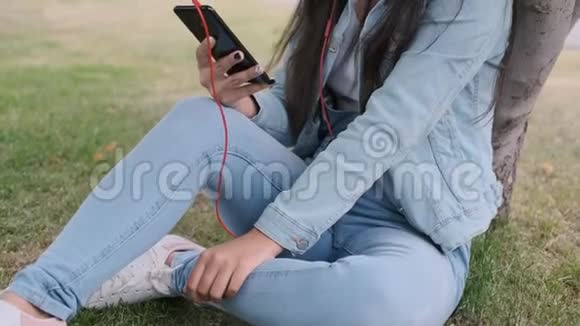 女孩坐在公园的一棵树旁听着耳机里的音乐特写视频的预览图