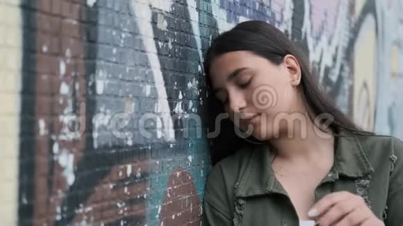 年轻漂亮的女孩站在墙边涂鸦摆姿势用手穿过头发看着摄像机视频的预览图