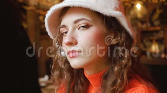 咖啡馆里迷人的漂亮女孩看起来很可爱年轻卷曲的绿眼睛女人戴着时髦的蓬松帽子和橙色毛衣视频的预览图