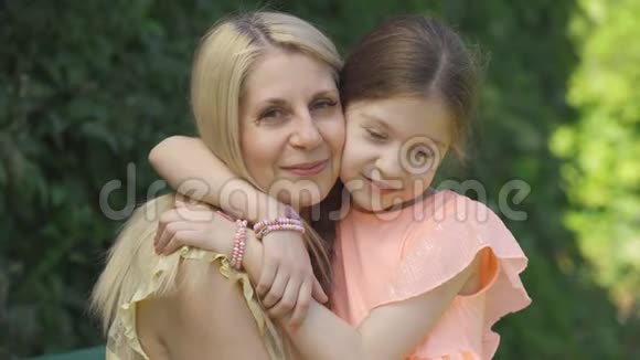 一幅金发母亲和她的小女儿拥抱在夏天公园看镜头的肖像家庭幸福美满视频的预览图