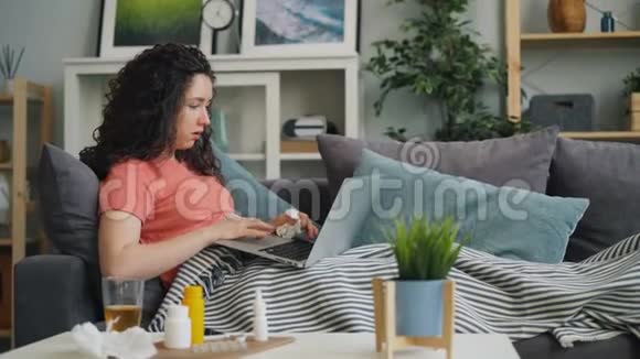 生病的年轻女士用手提电脑浏览沙发咳嗽摸鼻子视频的预览图