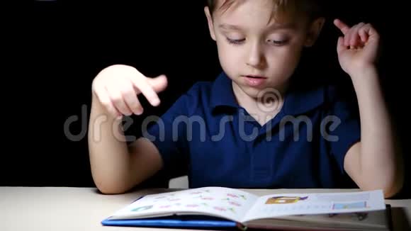 一个孩子晚上坐在一张灯点亮的桌子旁读一本书用手指指着书中的图画视频的预览图