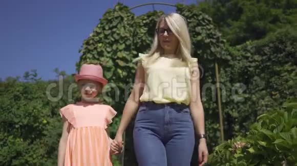 可爱的金发母亲和可爱的小女儿在夏天的花园里牵手散步家庭幸福妇女和女孩视频的预览图