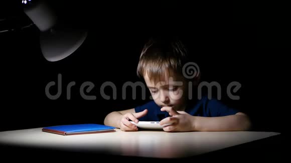 一个孩子晚上在家里小心翼翼地看着一幅漫画他用一部智能手机坐在一张灯点亮的桌子旁视频的预览图