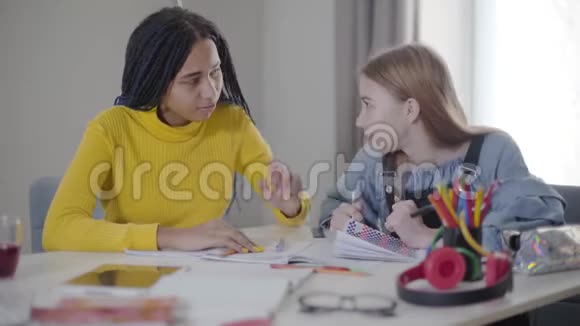 积极的白人少女与非裔美国朋友交谈两个学生在家一起做作业学习视频的预览图