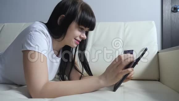 女孩用数字平板电脑制作网上销售购物概念黑发女人躺在沙发上看着网上商店通过支付视频的预览图