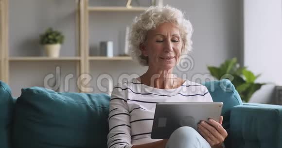 微笑的高级女性使用数码平板电脑在沙发上放松视频的预览图