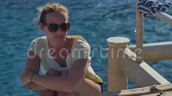 阳光明媚的一天戴太阳镜的年轻女孩坐在海边视频的预览图