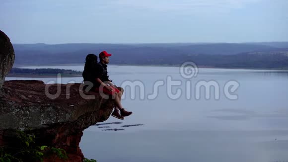 一个快乐的徒步旅行者坐在悬崖边上在一座岩山的山顶上举起双臂动作缓慢视频的预览图