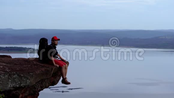 一位背着背包的旅行者坐在悬崖边在岩石山的山顶上摆动着腿视频的预览图