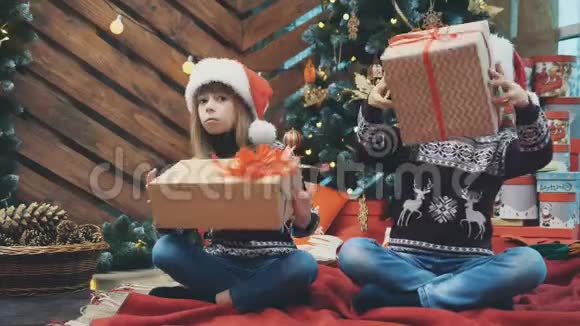 小双胞胎坐在圣诞树下摇晃着摆弄着礼物盒找出里面是什么视频的预览图