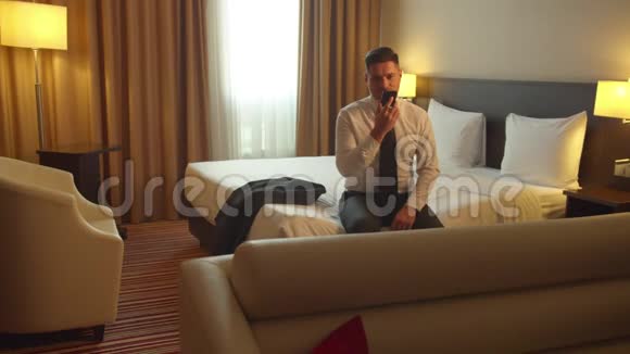 穿着衬衫和领带的男人坐在酒店的床上在电话中记录语音信息视频的预览图