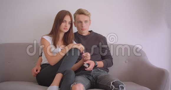 一对年轻可爱的白种人夫妇坐在室内沙发上看恐怖电影的特写镜头视频的预览图