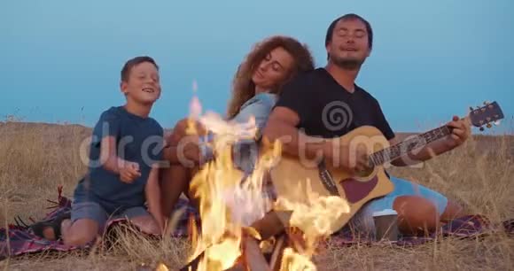 爸爸弹吉他妈妈和儿子在篝火旁放松家庭野餐特写镜头视频的预览图