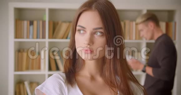 一张年轻漂亮的白人女性的特写照片她看着站在图书馆里的相机一个年轻的男性正在阅读视频的预览图