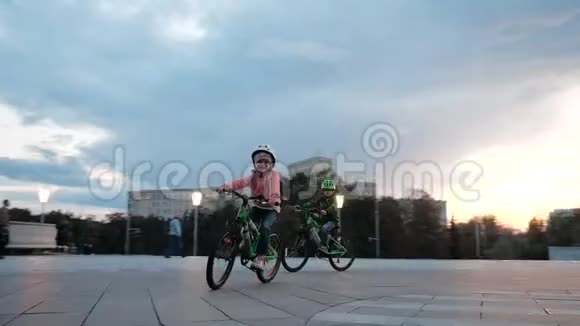 一个微笑的小女孩和男孩骑绿色自行车在一个城市的晚上在一个圆圈戴着头盔的小自行车视频的预览图
