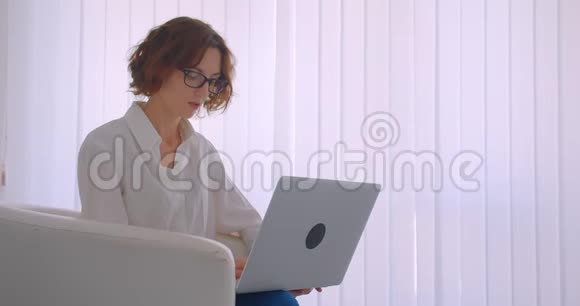 戴着眼镜的白种人成年女商人用笔记本电脑看镜头坐着的特写照片视频的预览图