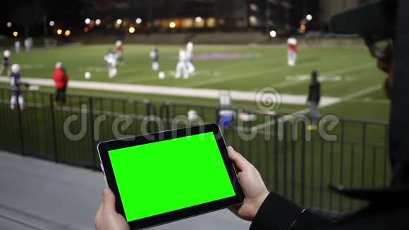 男子在V2露天看台的足球队练习赛上观看绿屏平板视频的预览图