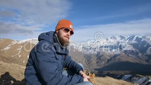 一张戴着太阳镜和帽子的留胡子旅行者的肖像坐在山峦背景下的岩石上拒绝视频的预览图