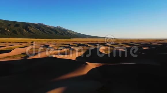 在明亮的蓝天下用绿色山脉拍摄黄色沙丘的空中镜头大沙丘国家公园视频的预览图