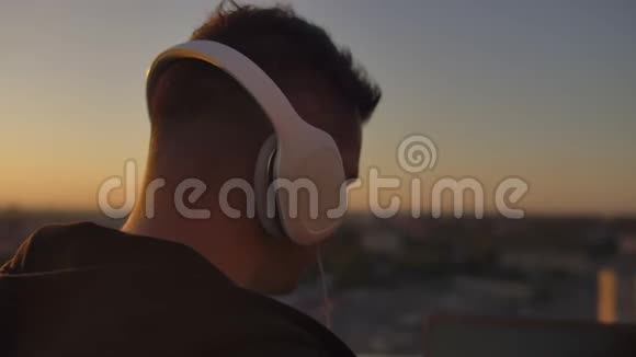 日落时分一个戴着耳机在屋顶上听音乐的人的特写镜头可以看到这座城市视频的预览图