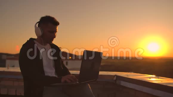 日落时分一个戴着耳机听音乐在建筑物屋顶上工作的人的后视镜视频的预览图